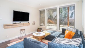 Un salon moderne doté d’un grand téléviseur à écran plat fixé au mur, d’un coin salon confortable avec un canapé et un pouf, et d’une grande fenêtre offrant une vue sur l’extérieur.