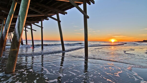 Un muelle de madera que se adentra en el océano, con el sol poniéndose al fondo.