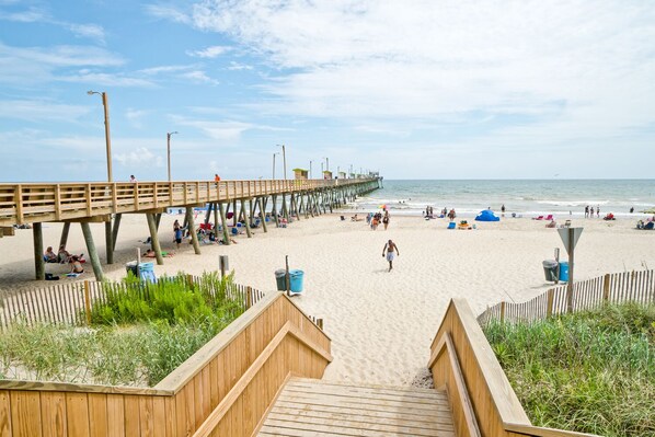 A wooden pier extending over a sandy beach with people walking and sitting.