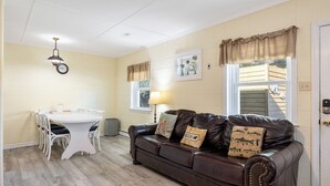 Una habitación acogedora con un sofá de cuero marrón, una mesa de comedor blanca con sillas, un reloj en la pared y una ventana con cortinas.
