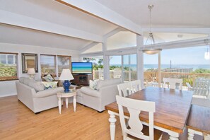 A spacious living room with a wooden dining table, white chairs, and a television. - Coral Palms Cottage, Oceanfront House in Emerald Isle, NC! (Emerald Isle)