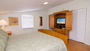 Quarto com cama, guarda-roupa de madeira, televisão, janela com persianas e uma obra de arte pendurada na parede.