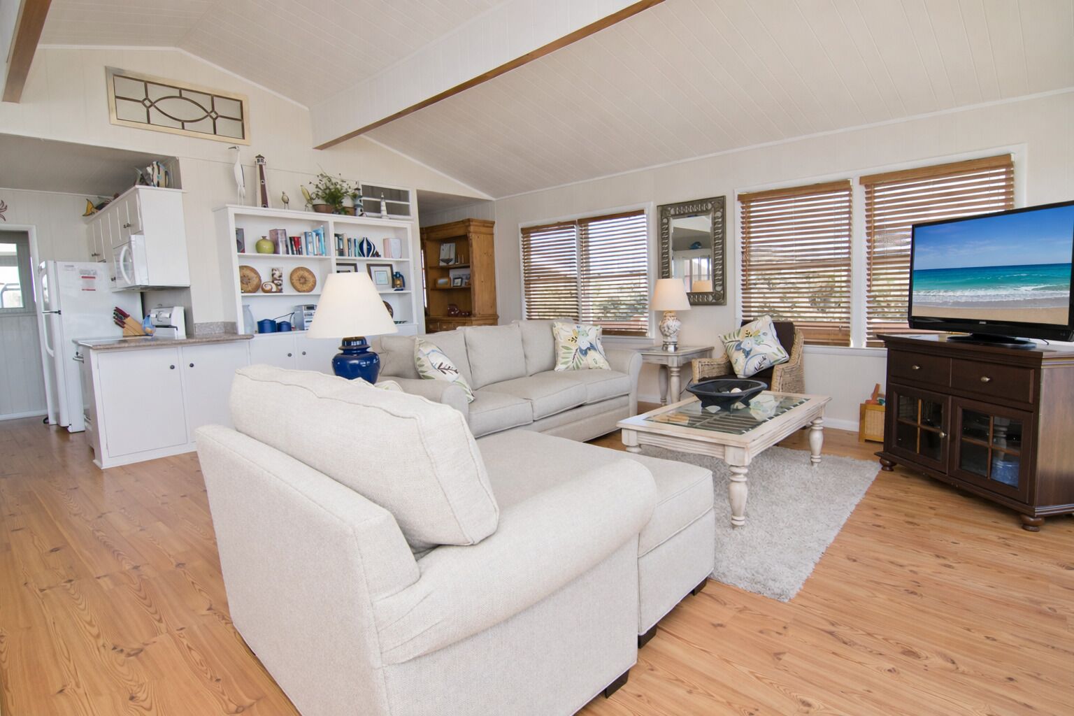 A spacious living room with a sofa, a coffee table, and a television. There is a kitchen area with cabinets and a refrigerator.