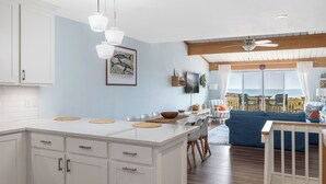 Cocina moderna con armarios blancos, una isla central, iluminación colgante y un área de comedor con sofá azul y piso de madera.