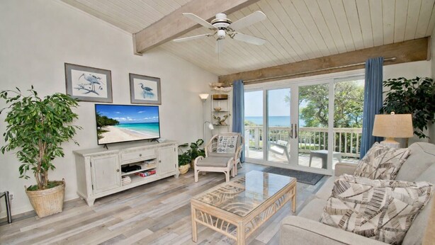 A spacious living room with a large TV, comfortable seating, a ceiling fan, and a view of the ocean through large sliding doors.