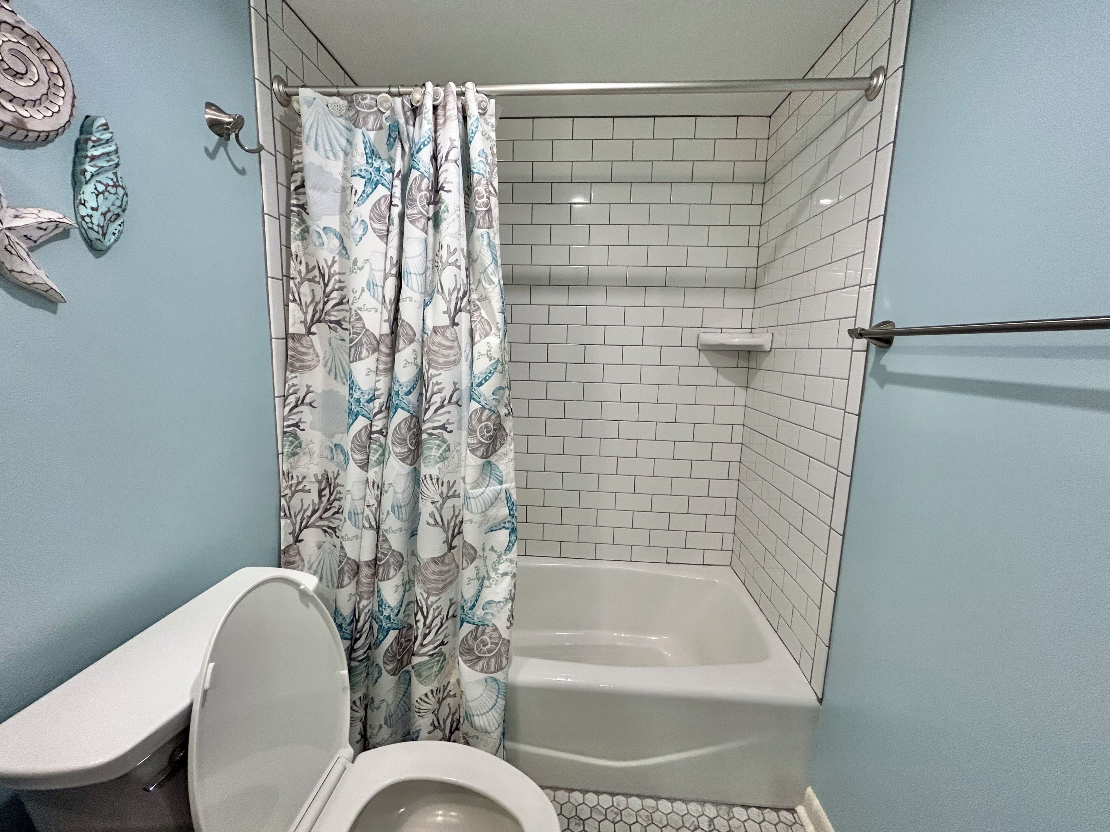 A bathroom with a toilet, shower, and bathtub, featuring a tiled wall and a curtain with a shell pattern.