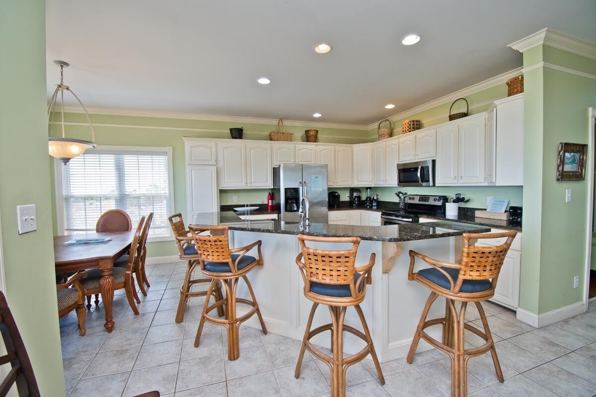 A kitchen with white cabinets, a central island with bar stools, a dining table with chairs, and a window with blinds.