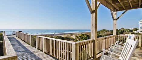 A wooden deck with a railing overlooking the ocean, white lounge chairs, and a covered area with a view of the beach.