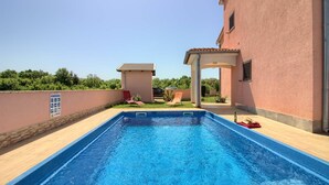 A rectangular swimming pool with a clear blue water surface, surrounded by a wooden deck. There are two red lounge chairs and a small covered seating area with a table. The pool is adjacent to a building with a pink exterior and a tiled roof. - Villa Mary (Istria - Liznjan) (Liznjan)