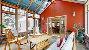 A sunroom with large windows, a wooden bench, a dining area, and potted plants.