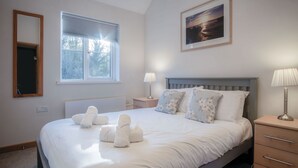 Un lit bien fait, avec du linge de lit et des serviettes blanches, une table de chevet avec une lampe, une fenêtre avec des stores et un tableau encadré au mur.