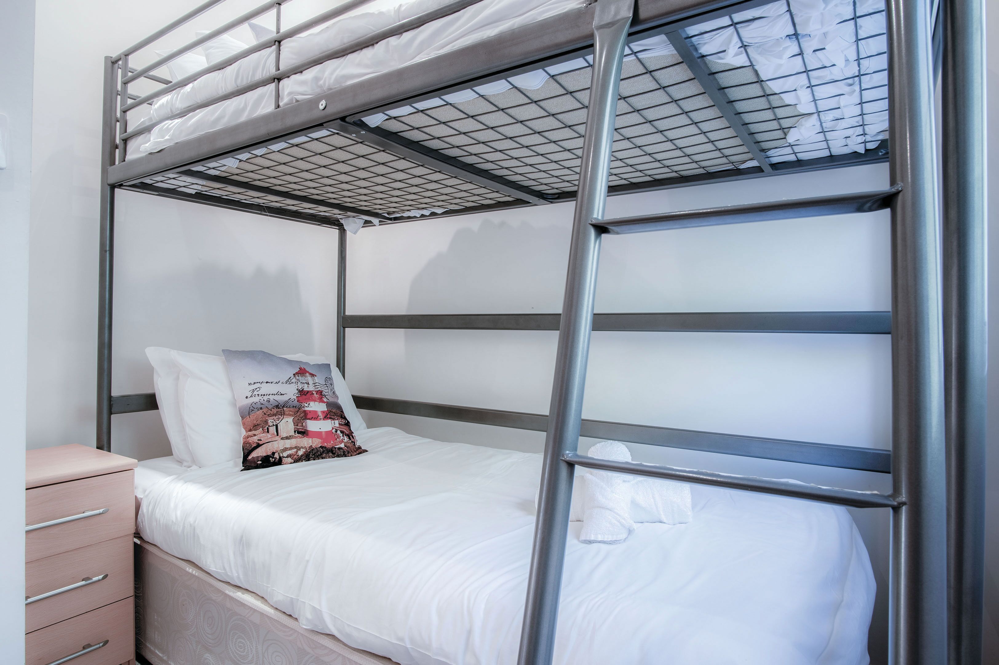 Una cama litera con estructura metálica, ropa de cama blanca y un almohadón con diseño de faro.