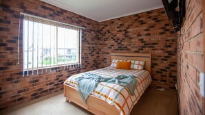 A bedroom with a brick wall, a wooden bed, and a window with blinds. - Beachview 2 - Scenic Drive 33 (Bermagui)
