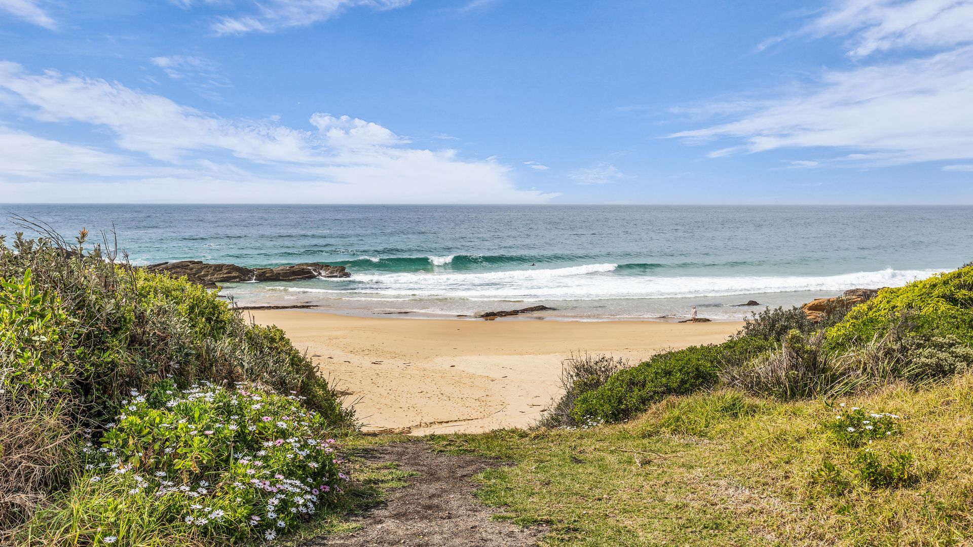 A beach with a clear view of the ocean, a sandy path leading to the shore, and surrounding greenery.