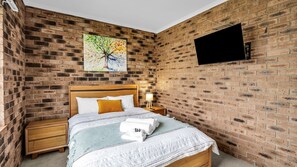 A bedroom with a brick wall, a bed with white bedding, a wooden headboard, a nightstand with a lamp, and a painting of a tree.