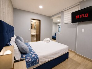A hotel room with a bed, a bedside table, a TV mounted on the wall, and a bathroom visible through an open door. - Double/Triple Suite 18-103 (Bogotá)