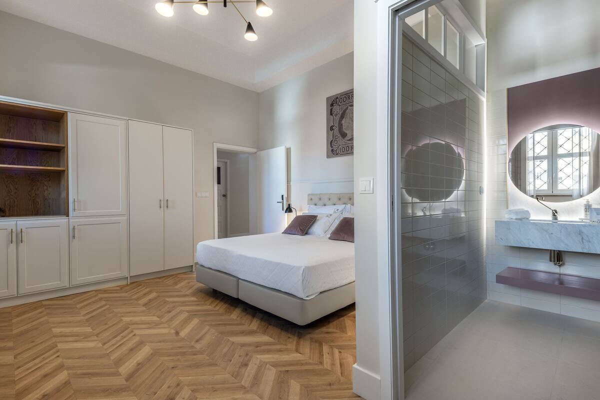 A modern bedroom with a large bed, built-in wardrobe, and a glass-enclosed bathroom with a vanity and mirror.