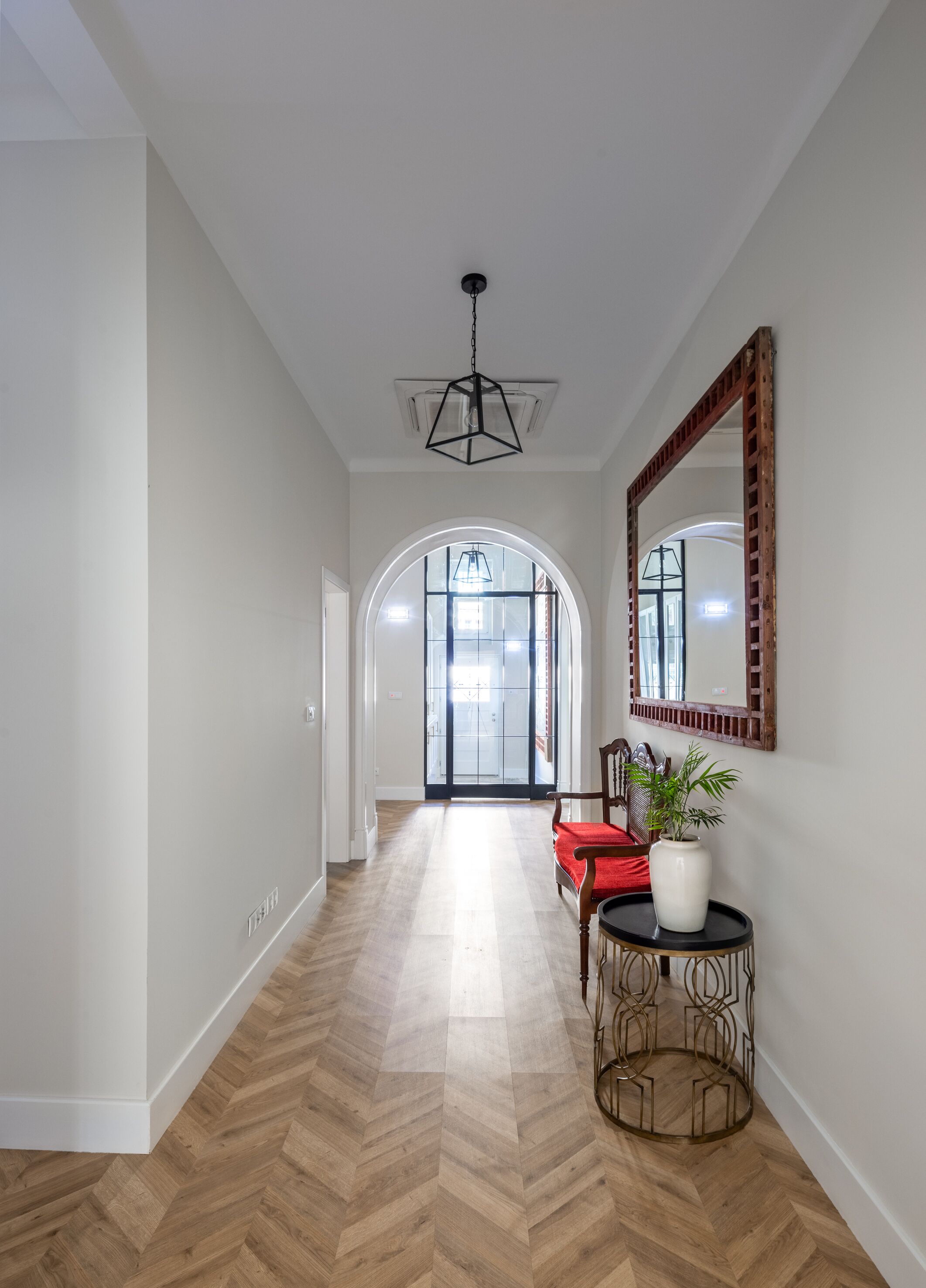 Un pasillo con suelo de espiga, una silla de madera con cojín rojo, un jarrón blanco con planta y un espejo en la pared.
