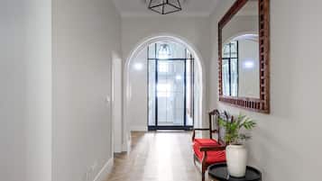 Un pasillo con suelo de espiga, una silla de madera con cojín rojo, un jarrón blanco con planta y un espejo en la pared.