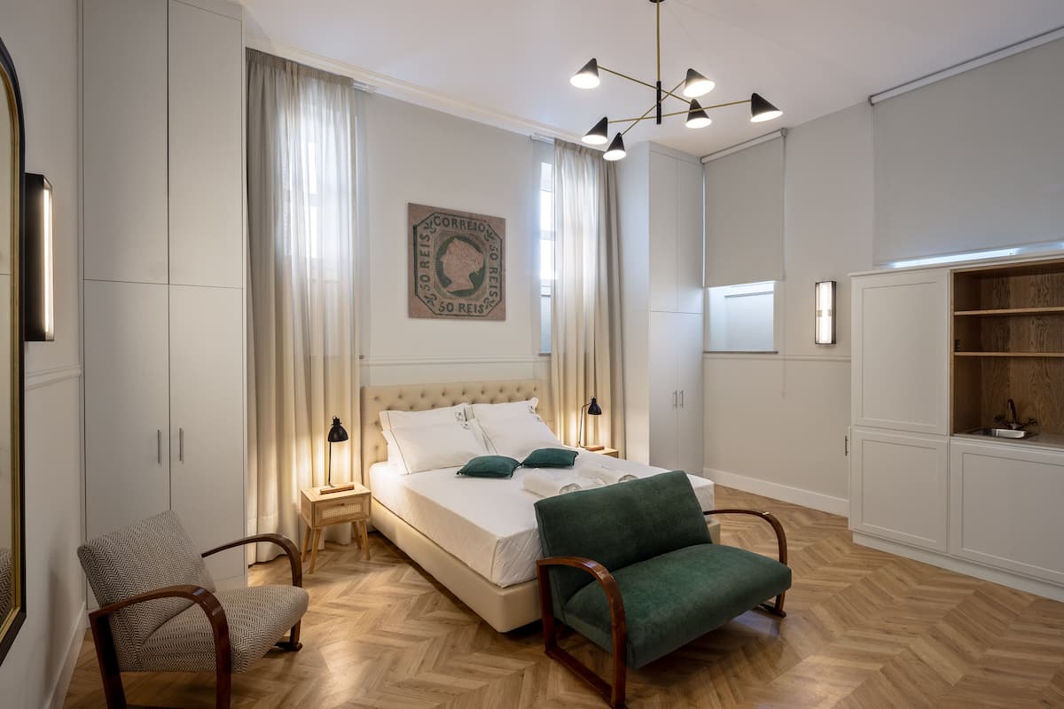 A bedroom with a bed, two chairs, a nightstand, a wall-mounted lamp, and a painting.