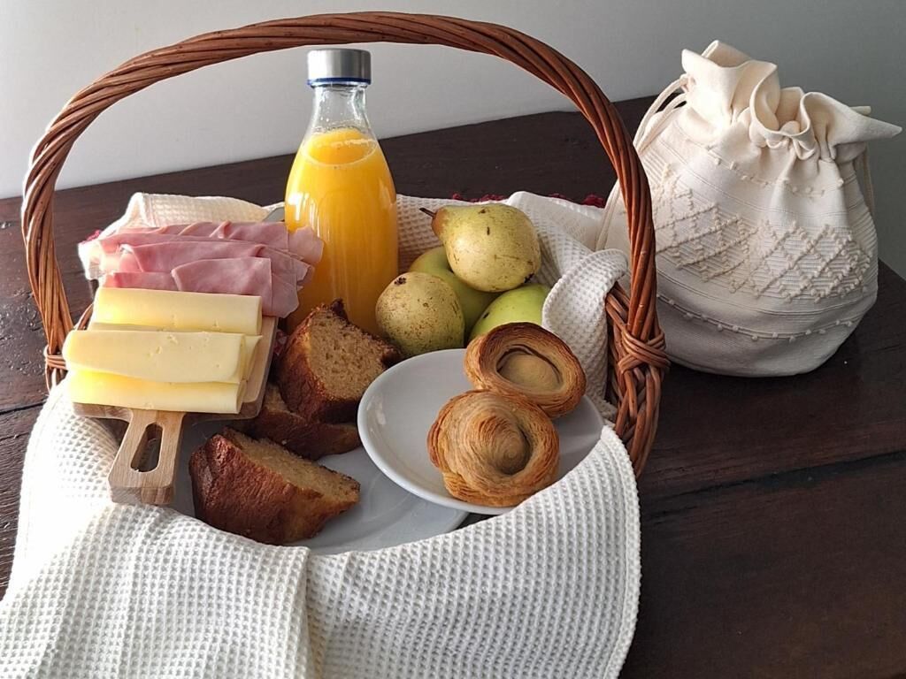 Una cesta de mimbre con artículos de desayuno como queso, jamón, pan, bollería y una botella de zumo de naranja.