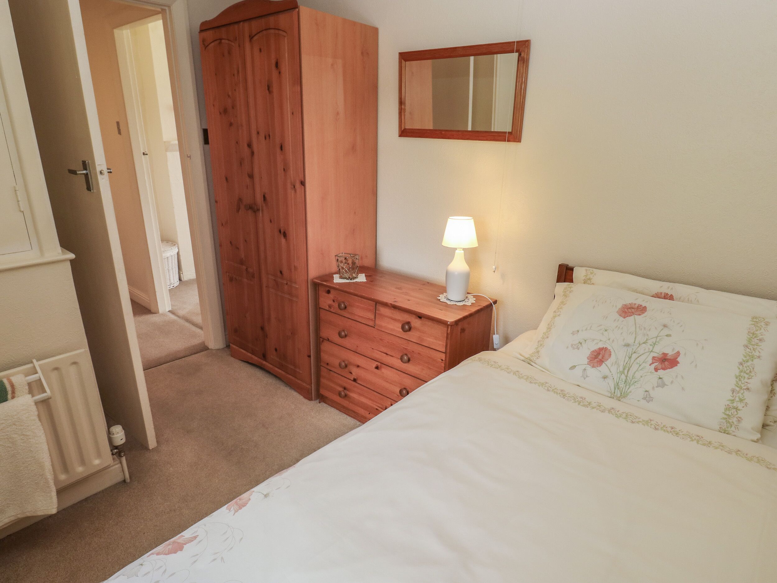 A bedroom with a wooden wardrobe, a bedside table with a lamp, and a bed with floral bedding.