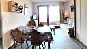 Een kleine kamer met een eettafel, stoelen, een bed, een televisie en een balkon met gordijnen.