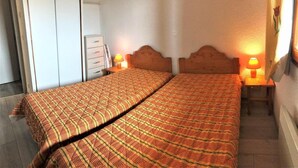 Een tweepersoonsbed met een geruite sprei, twee bedlampjes, een houten hoofdbord, een kleine tafel met lamp en een garderobe.
