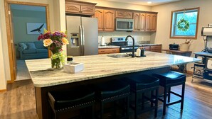 A kitchen with a large island, wooden cabinets, a stainless steel refrigerator, a microwave, and a stove. There are three bar stools at the island.
