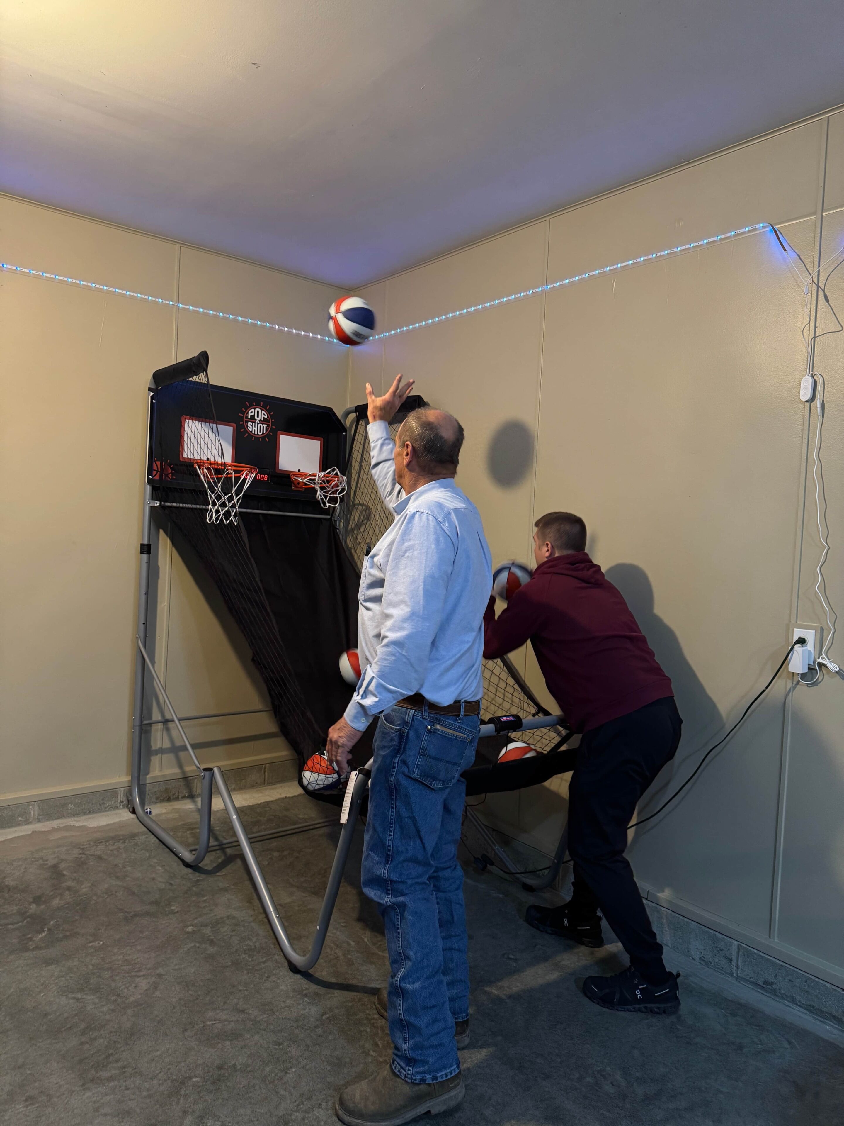 A room with a basketball hoop, a person playing, and another person assisting.