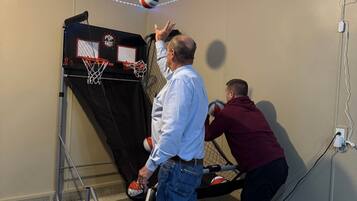A room with a basketball hoop, a person playing, and another person assisting.