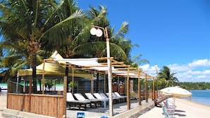 Un bar situé en bord de mer, doté de chaises longues et de parasols, entouré de palmiers et offrant une vue dégagée sur l’océan.