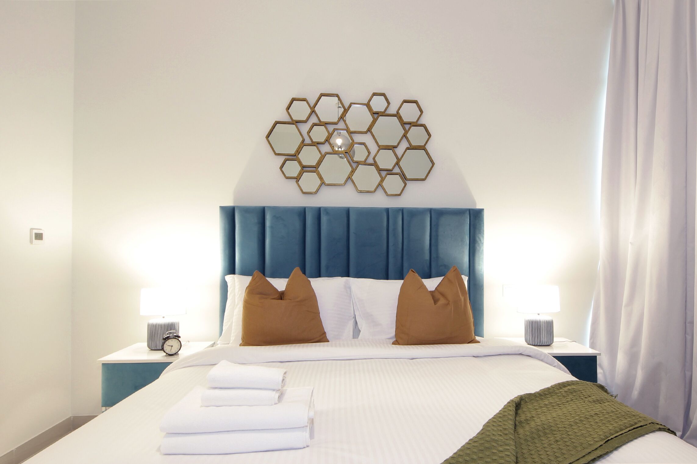 Cama bem arrumada com lençóis brancos, duas almofadas marrons e um cobertor verde. Há duas mesas de cabeceira com abajures e um espelho hexagonal fixado na parede.