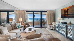 A spacious living room with a large sofa, a coffee table, and a television. The room has a large window with a view of the ocean. There is a balcony with a table and chairs. - 🚢Beachfront Complex-Private Beach-Incredible Gulf Views-Edgewater #1007🚢 (Miramar Beach)
