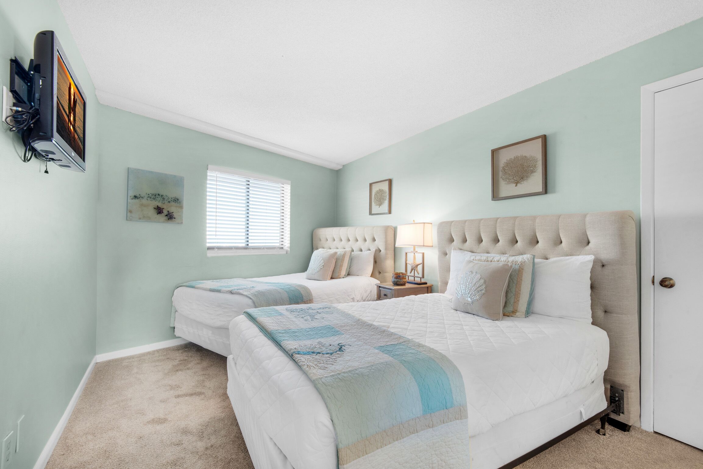 A bedroom with two beds, a television mounted on the wall, and a window with blinds.