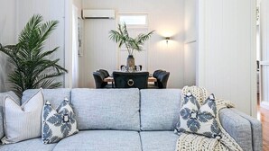 A living room with a grey sofa, two patterned pillows, a large green plant, and a dining area with a table and chairs. - Central Haven Milton 4BR Sleeps 10 (Milton)