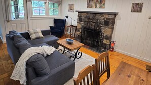 A living room with a sofa, a blue armchair, a fireplace, and a window with curtains. - Butternut Adirondack Cabin. (Johnsburg)