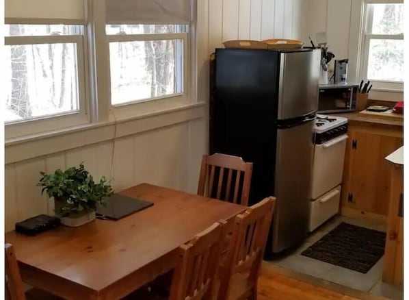 Une cuisine compacte équipée d’un réfrigérateur, d’une cuisinière et d’un four à micro-ondes. On y trouve une table à manger en bois avec des chaises et une plante en pot.