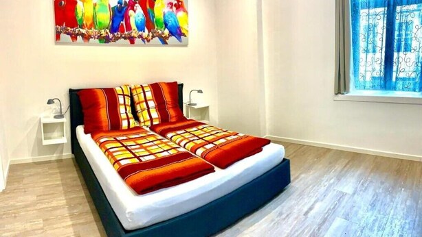 Un dormitori modern amb un llit gran, una obra d'art colorida sobre el llit i una finestra amb cortines.