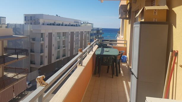 A balcony with a table and chairs, a refrigerator, and a view of the sea and buildings. - Bilocale Ariosto Vista Mare Laterale (Gallipoli)