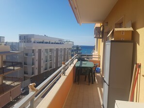 Un balcón con una mesa y sillas, un refrigerador y vistas al mar y edificios.