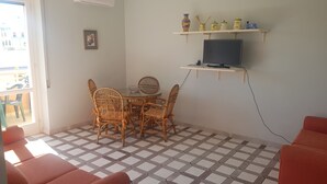 A room with a dining table and chairs, a TV on a shelf, and a patterned floor. - Bilocale Ariosto Vista Mare Laterale (Gallipoli)