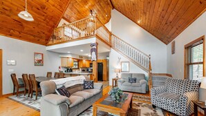 A spacious living area with a high ceiling, wooden flooring, a dining table with chairs, a sofa, a patterned armchair, and a staircase leading to an upper level.