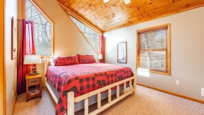 A bedroom with a wooden ceiling, a bed with a red and black checkered bedspread, a nightstand with a lamp, and a window with blinds.