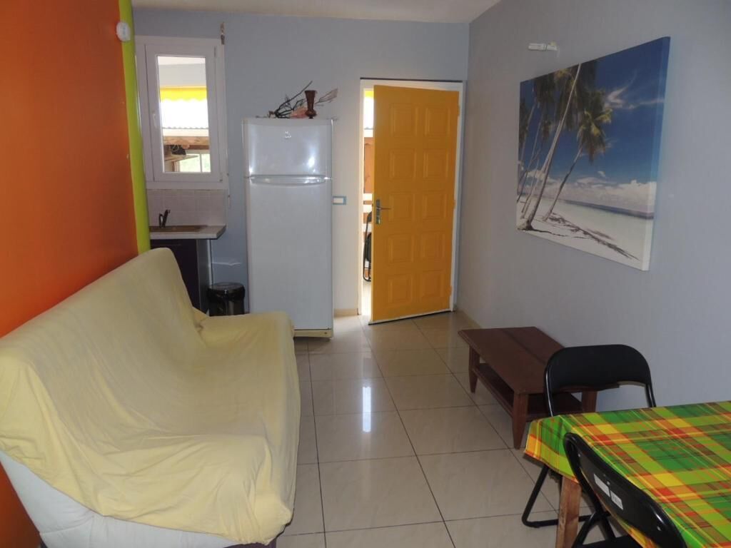 A room with a white armchair, a small table with a checkered tablecloth, a refrigerator, a door leading to another room, and a painting of a beach scene.