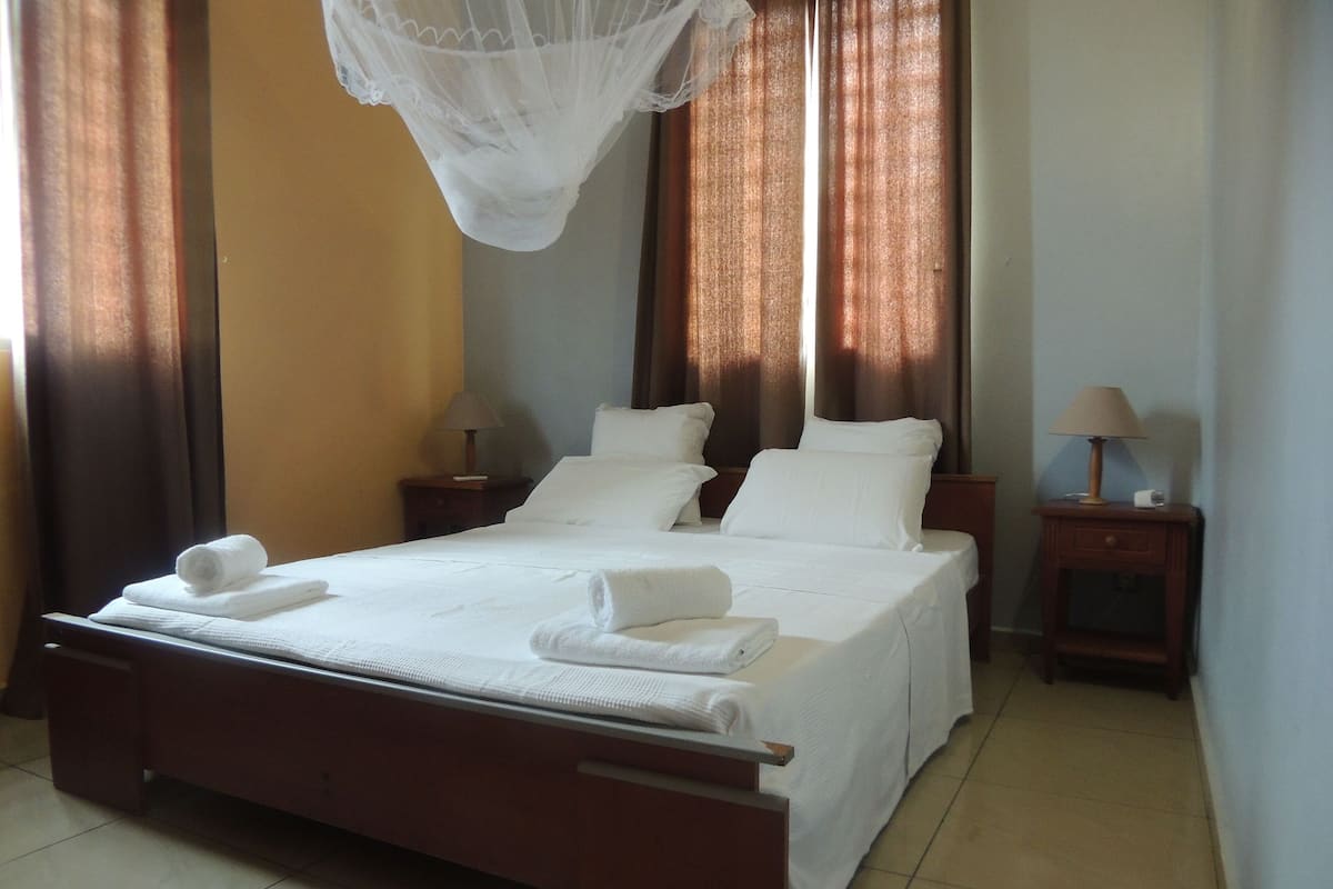 A neatly made bed with white linens and towels, a bedside table with a lamp, and a window with curtains.