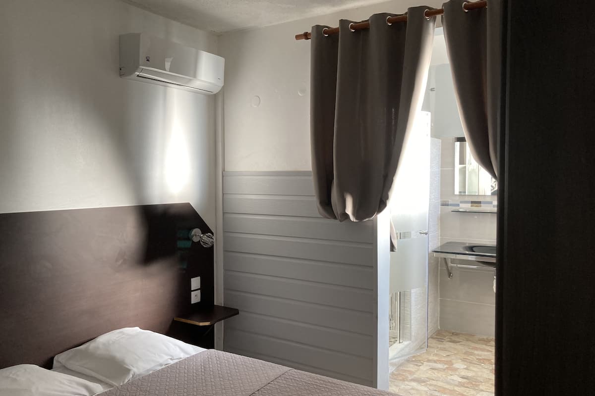 A hotel room with a bed, an air conditioning unit, a small table with a lamp, and a bathroom visible through a door.