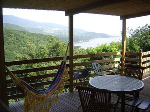 A wooden deck with a hammock, chairs, and a table overlooking a mountainous landscape and a body of water. - Hotel Gîtes Couleur Caraïbes (Bouillante)