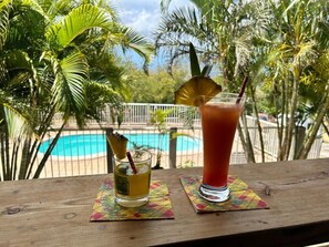 Two drinks on a wooden table with a pool and palm trees in the background. - Hotel Gîtes Couleur Caraïbes (Bouillante)