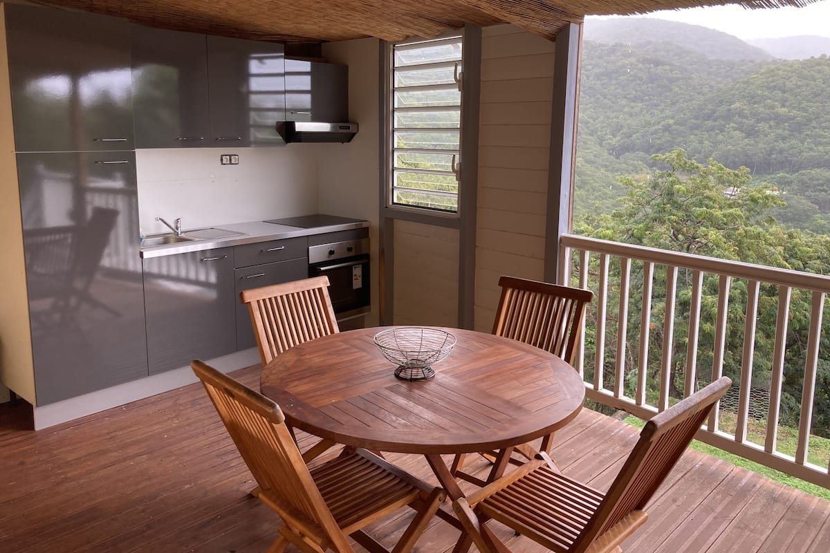 A wooden deck with a round table and chairs, a kitchen with grey cabinets, and a view of lush green hills.